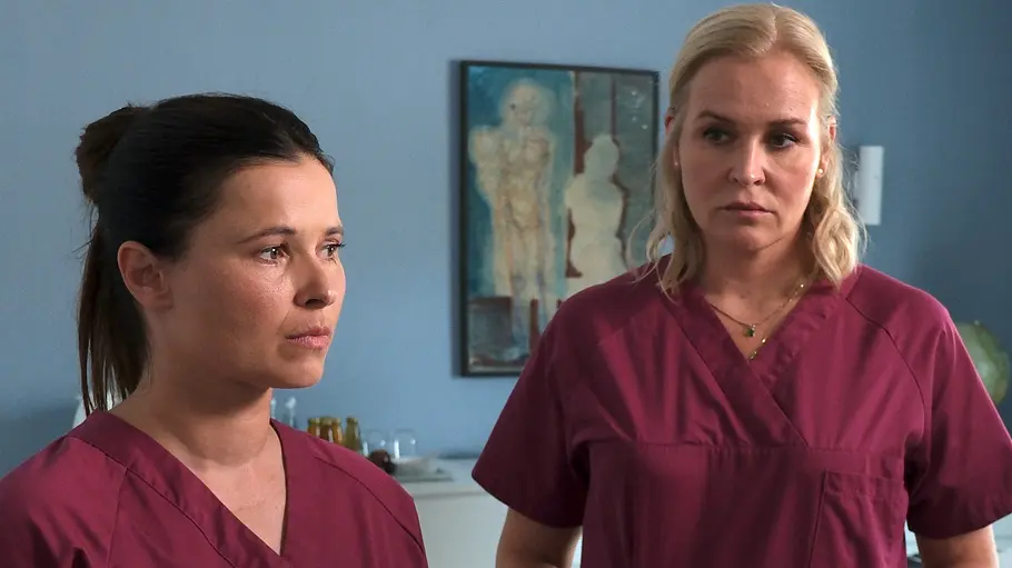 Als Lena Lorenz (Judith Hoersch, r.) ihre Kollegin Gesa Hahn (Anna Fischer) bezüglich des Geburtsverlaufs bei Fine Kleinert zur Rede stellt, reagiert Gesa beleidigt. Lena Lorenz - Auf Augenhöhe: In einem Behandlungszimmer steht Lena Lorenz (Judith Hoersch) mit sehr ernstem Gesichtsausdruck und blickt Hebamme Gesa (Anna Fischer) an, die mit starrem Blick geradeaus schaut. Im Hintergrund hängt ein Gemälde an einer hellblau gestrichenen Wand.