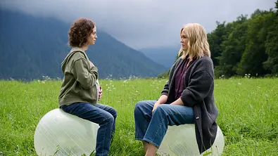 Auf einer Wiese in den Bergen sitzen sich Sina Schumacher (Fanny Altenburger) und Lena Lorenz (Judith Hoersch) auf großen Gymnastikbällen gegenüber. Sina legt eine Hand auf ihren Bauch, die andere auf ihre Brust Beide blicken sich ernst an. - Foto: ZDF/ WEHNER_WALTER