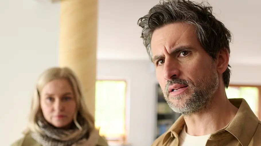 Tom Beck schaut überrascht und leicht besorgt in die Kamera, während im Hintergrund Lena Lorenz unscharf zu sehen ist, die ebenfalls ernst wirkt; beide befinden sich in einem hellen Innenraum.