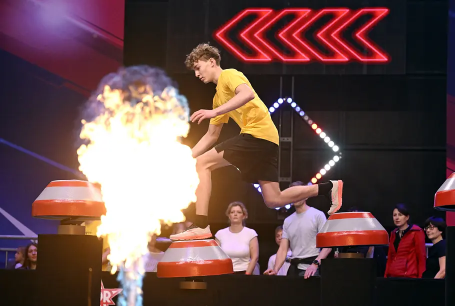 Leo Rothermel ist Teilnehmer bei Ninja Warrior Germany 2026 Leo Rothermel im gelben T-Shirt springt über rot-silberne Podeste. Im Vordergrund lodert eine Flamme, dahinter stehen Zuschauer.