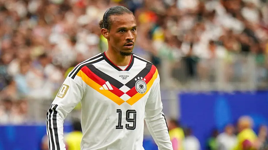 Ein deutscher Nationalspieler mit zurückgebundenen Haaren trägt während eines Länderspiels das weiße DFB-Trikot mit der Rückennummer 19 und blickt konzentriert auf das Spielgeschehen. Im Hintergrund sind die Zuschauer im Stadion unscharf zu sehen.