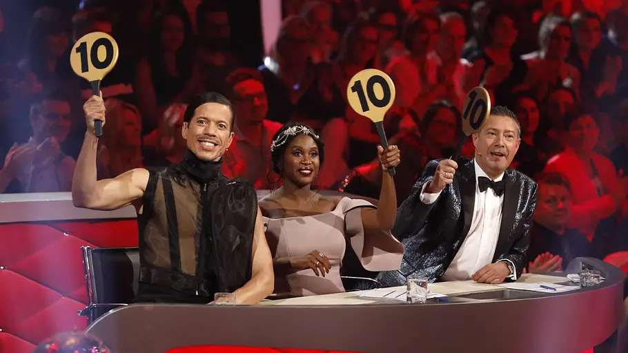 Let's Dance Jury Drei Jury-Mitglieder sitzen an einem rot beleuchteten Jury-Pult in einer TV-Tanzshow und halten jeweils eine Bewertungstafel mit der Zahl 10 hoch, während im Hintergrund ein jubelndes Publikum zu sehen ist.