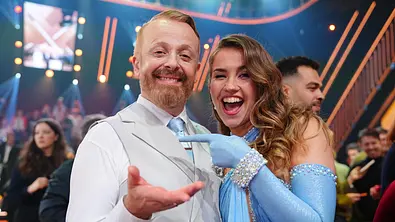 Ein fröhliches Tanzpaar steht Arm in Arm auf einer bunt beleuchteten TV‑Bühne und lächelt in die Kamera, während im Hintergrund Studiogäste zu sehen sind. - Foto: RTL / Stefan Gregorowius