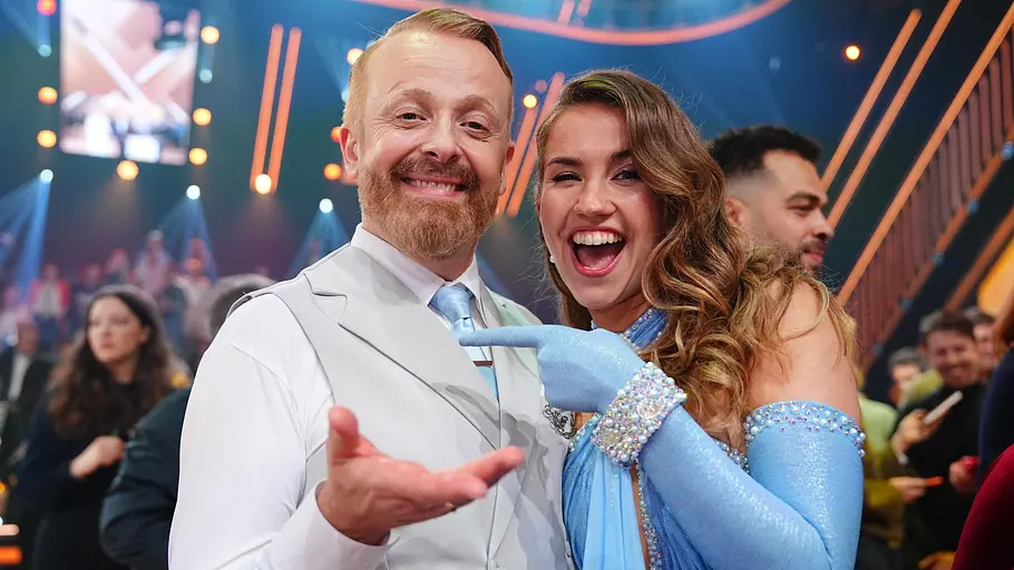 Let's Dance Paar: Ross Anthony und Mariia Maksina Ein fröhliches Tanzpaar steht Arm in Arm auf einer bunt beleuchteten TV‑Bühne und lächelt in die Kamera, während im Hintergrund Studiogäste zu sehen sind.