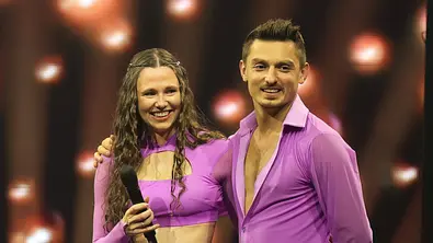 Bike-Influencerin Ann-Kathrin Bendixen und Tänzer Zsolt Sandor Cseke in lilafarbenen Tanz-Outfits stehen lächelnd nebeneinander auf einer Bühne mit Lichterhintergrund; die Frau hält ein Mikrofon in der Hand. - Foto: IMAGO / Bildagentur Monn