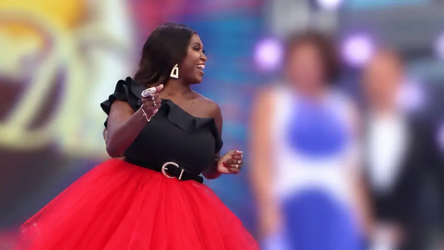Lets-Dance-Wechsel Motsi Mabuses Kollegin Motsi Mabuse im ausladenden roten Rock neben ihren Jury-Kollegen von „Strictly Come Daning“. Diese sind verschwommen dargestellt, genau wie der Hintergrund.