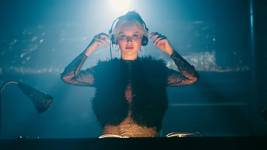 Linda Bierwirth (Jule Hermann) am DJ-Pult bei ihrem großen Auftritt. München Beats - Folge 4: Linda Bierwirth (Jule Hermann) steht hinter einem DJ-Pult und blickt in die Kamera. Sie steht vor dunklem Hintergrund, trägt Kopfhörer und wird von einer Leuchte, die über ihr hängt, angestrahlt.