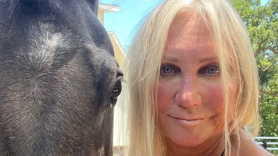 Linda Hogan im Freien, die neben einem schwarzen Pferd steht; beide blicken in die Kamera, im Hintergrund sind Bäume und ein Haus unter blauem Himmel zu sehen.