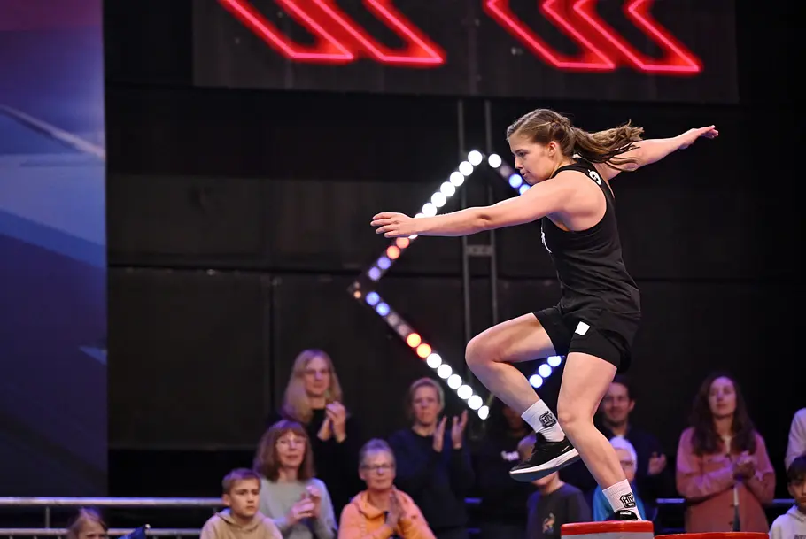 Lotta Kokemohr ist Teilnehmer bei Ninja Warrior Germany 2026 Lotta Kokemohr balanciert in schwarzer Sportkleidung mit ausgestreckten Armen auf einem Hindernis. Im Hintergrund klatscht Publikum.
