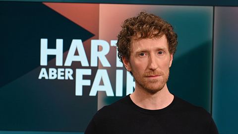 Louis Klamroth steht in einem schwarzen Oberteil vor dem „Hart aber fair“-Logo. - Foto: IMAGO / Horst Galuschka