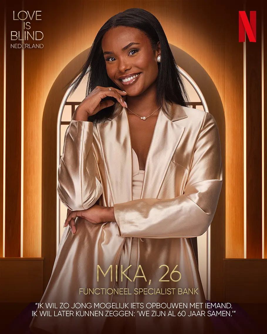 Love is Blind: Netherlands-Kandidat Mika Alter, Beruf Mika lächelt im champagnerfarbenen Satinblazer mit einer Hand am Kinn. Neben ihr stehen „Love is Blind Nederland“ und das Netflix-Logo.