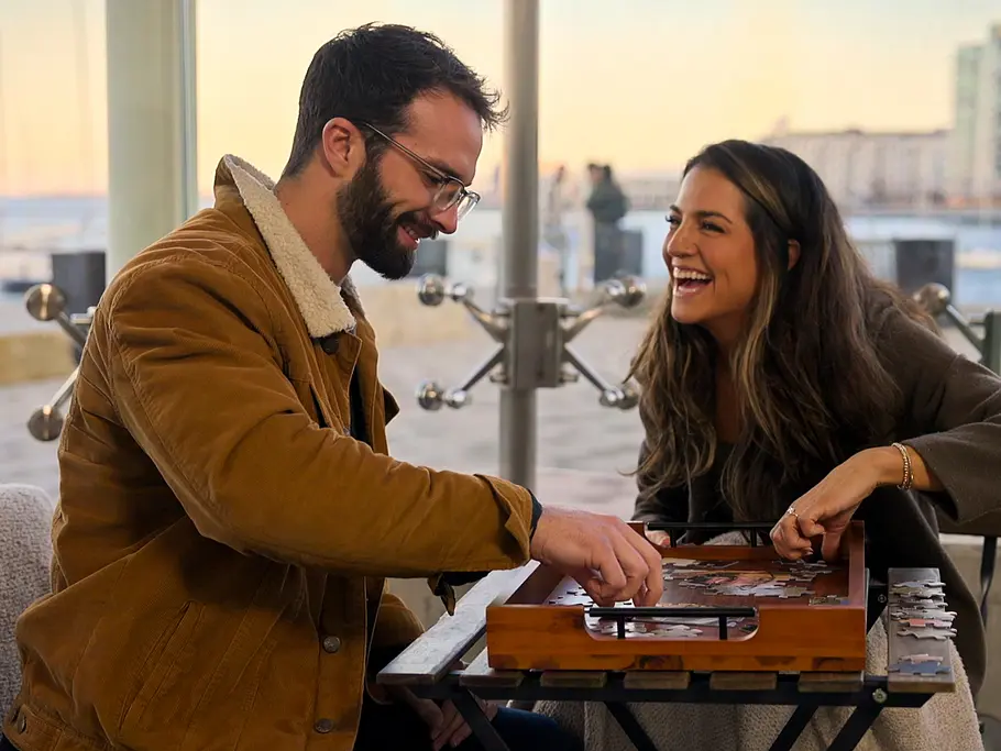 Sind Sarah und John noch zusammen? „Love is Blind:Boston“: Sarah und John sitzen lachend an einem kleinen Tisch mit einem Puzzle. Hinter großen Fenstern liegt eine Uferpromenade.