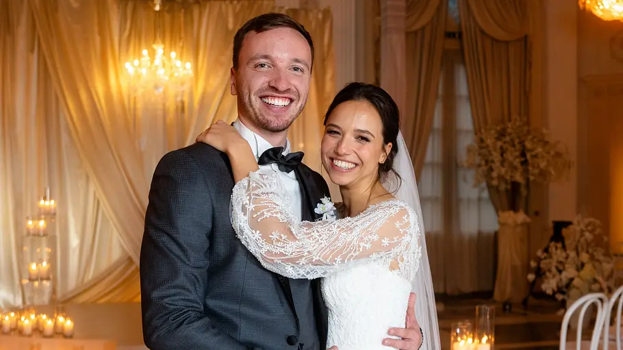 Ilias und Alina an ihrem Hochzeitstag. Alina und Ilias stehen Arm in Arm in eleganter Hochzeitskleidung in einem prunkvoll dekorierten Saal mit vielen Kerzen und Kronleuchtern. Beide lächeln glücklich in die Kamera.