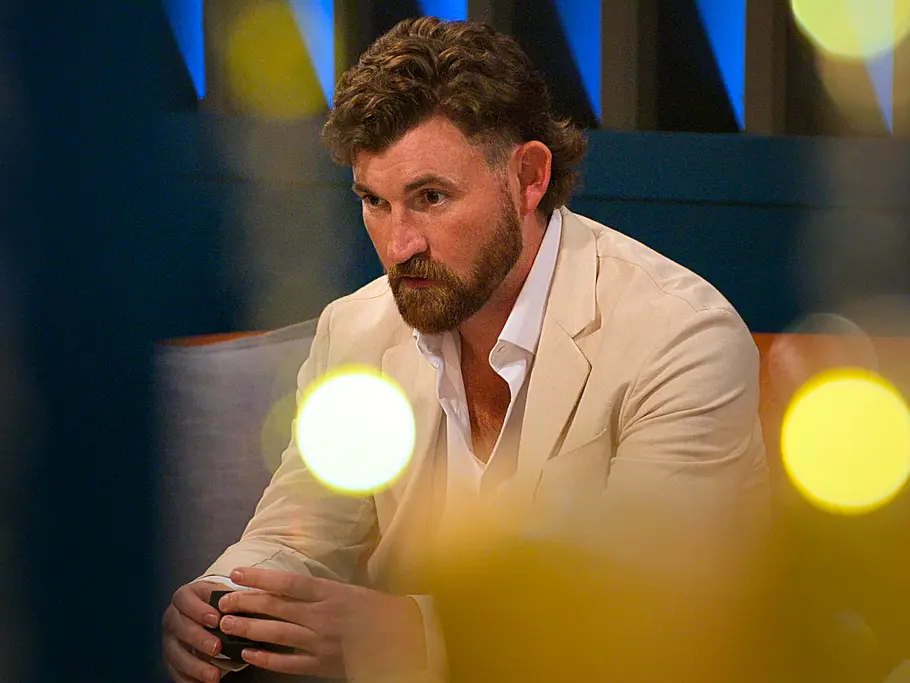 Blake Carver sitzt im hellen Anzug nach vorn gebeugt auf einem Sofa. Unscharfe gelbe Lichter liegen im Vordergrund.