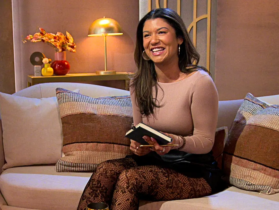Diana Perrone sitzt lächelnd mit einem Buch auf einem hellen Sofa. Vor ihr steht ein Polsterhocker mit einem goldenen Becher.