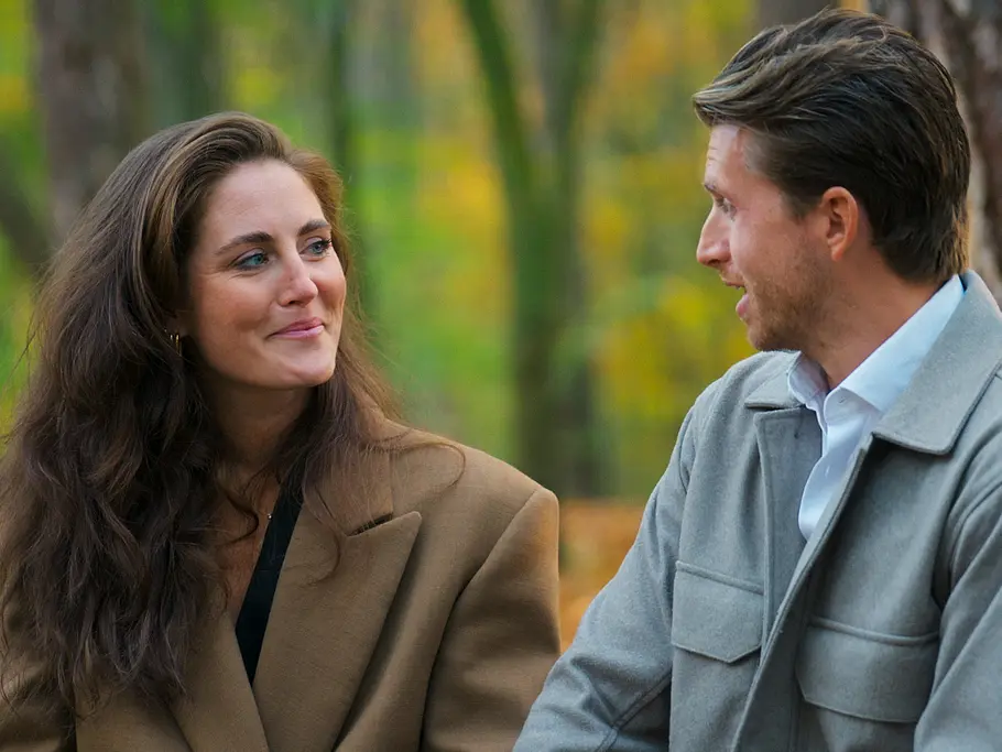 Mirella im braunen Mantel und Gino in einer grauen Jacke sitzen bei „Love is Blind: Netherlands“ für ein klärendes Gespräch im herbstlichen Wald und schauen einander an.