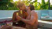 Ein Love is Blind: Nethterlands-Paar küsst sich mit goldenen Sektkelchen in einem Becken voller Rosenblätter. Dahinter stehen Liegen und Bananenpflanzen. - Foto: Netflix