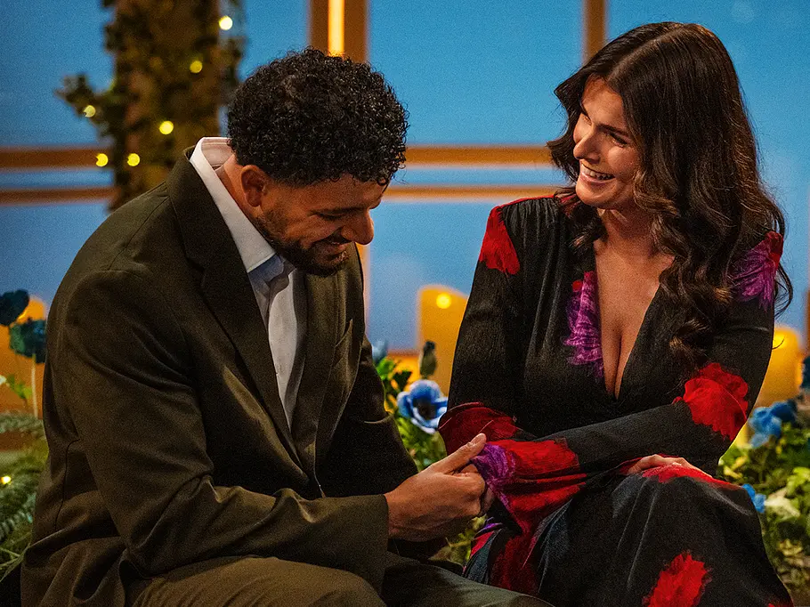 Yasemin und Roël sitzen beim „Love is Blind: Netherlands“-Reveal lächelnd auf einer blauen Bank und halten Händchen, umgeben von Blumen und leuchtenden Kerzen.