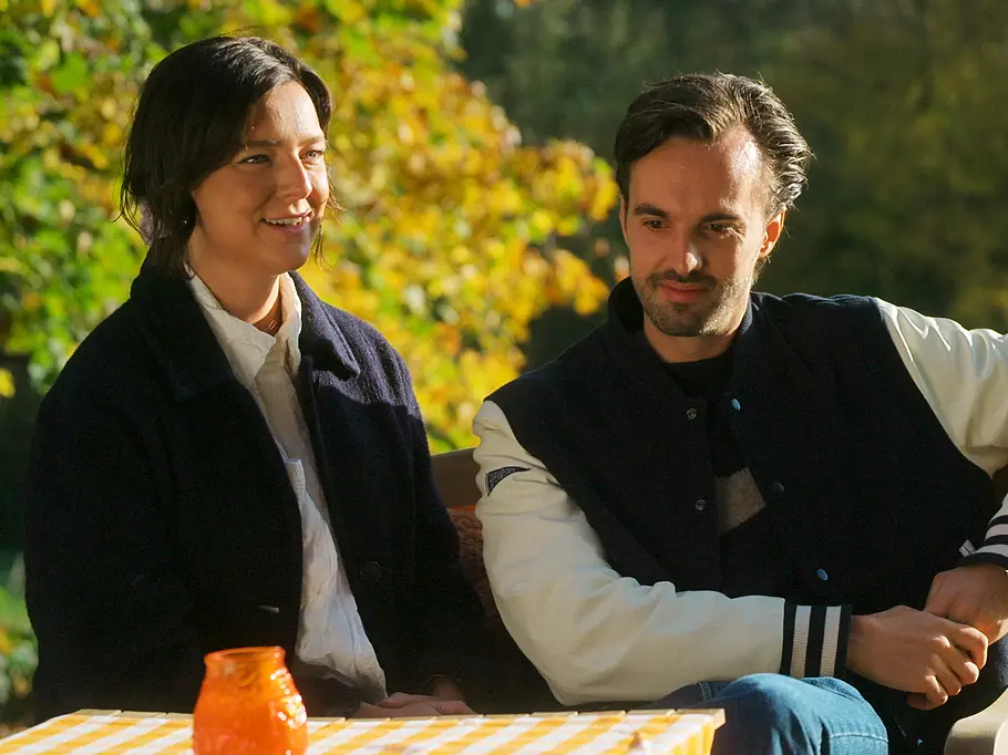 Meinou und Marc sitzen bei Love is Blind: Netherlands im Freien. Auf dem Tisch mit gelb-weiß karierter Decke steht ein orangefarbenes Glas.