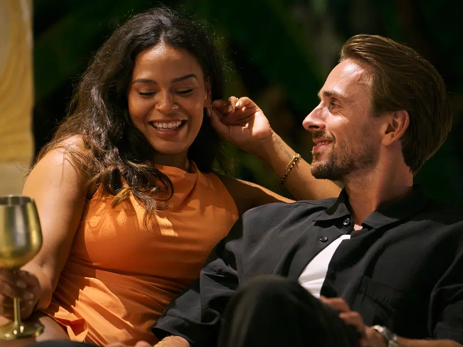 Jaslynn sitzt bei Love is Blind: Netherlands im orangefarbenen Kleid neben Ludo, sie hält einen goldfarbenen Kelch, beide lächeln.