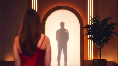 Love is Blind-Szene: Eine Frau im roten Kleid blickt auf eine Silhouette hinter einer beleuchteten Rundbogentür. Rechts steht eine Topfpflanze. - Foto: Paul Huttemann/Netflix