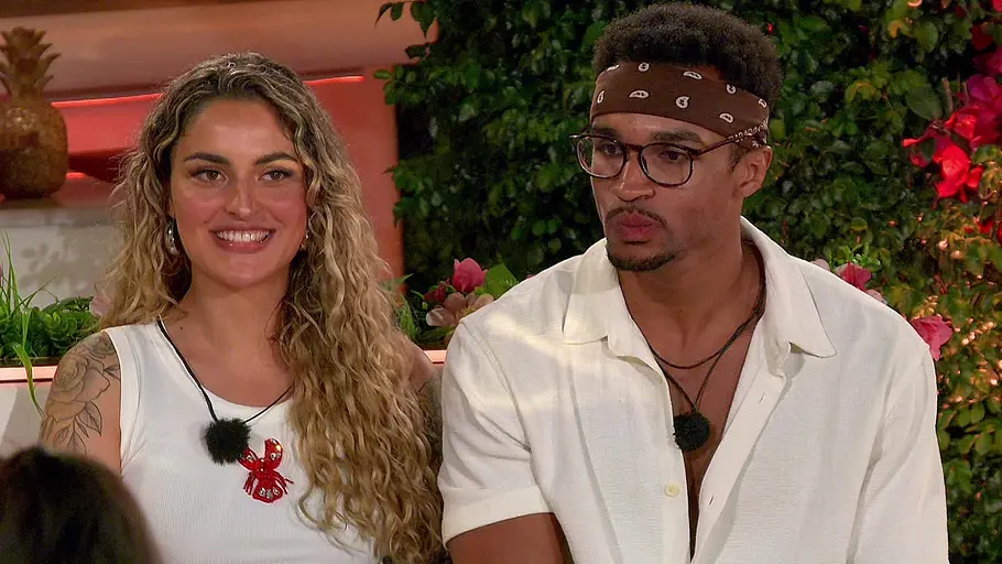 Chiaras Love Island VIP Chiara mit langen Locken sitzt neben David mit Brille und Bandana. Beide tragen Weiß; hinter ihnen sind Pflanzen.