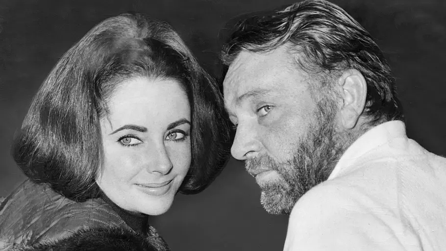 Elizabeth Taylor und Richard Burton blicken auf einer Schwarz-Weiß-Aufnahme über die Schulter in die Kamera und posieren eng nebeneinander.