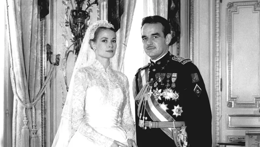 Grace Kelly steht in einem langen Brautkleid neben Fürst Rainier in Uniform in einem prunkvollen Saal, beide posieren für ein Hochzeitsfoto.