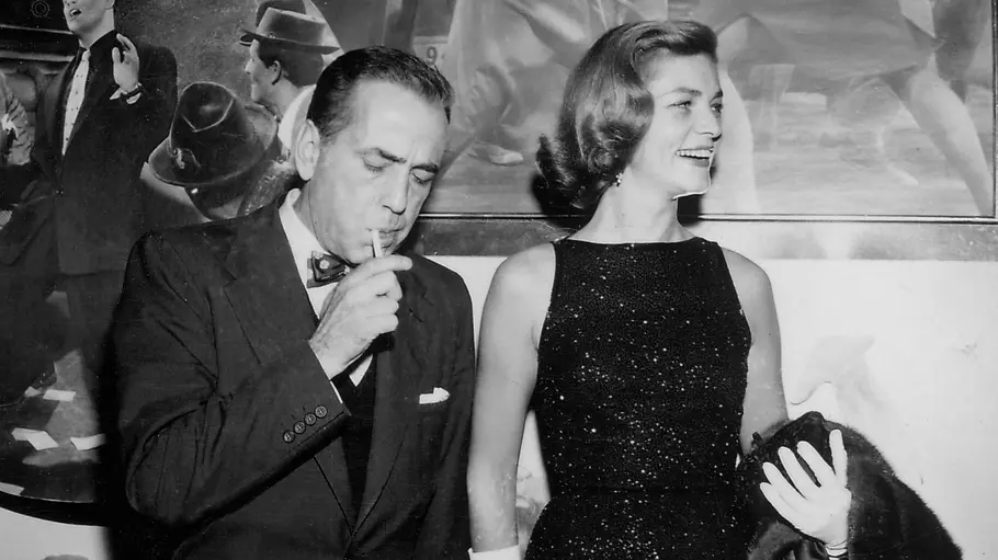 Humphrey Bogart steht neben Lauren Bacall auf einer Schwarz-Weiß-Aufnahme, während er raucht und sie in einem eleganten Abendkleid lächelnd zur Seite blickt.