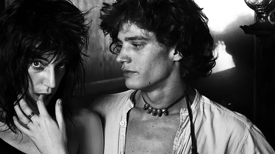 Patti Smith und Robert Mapplethorpe posieren auf einer Schwarz-Weiß-Aufnahme eng nebeneinander, während er sie ansieht und sie mit der Hand am Mund in die Kamera blickt.