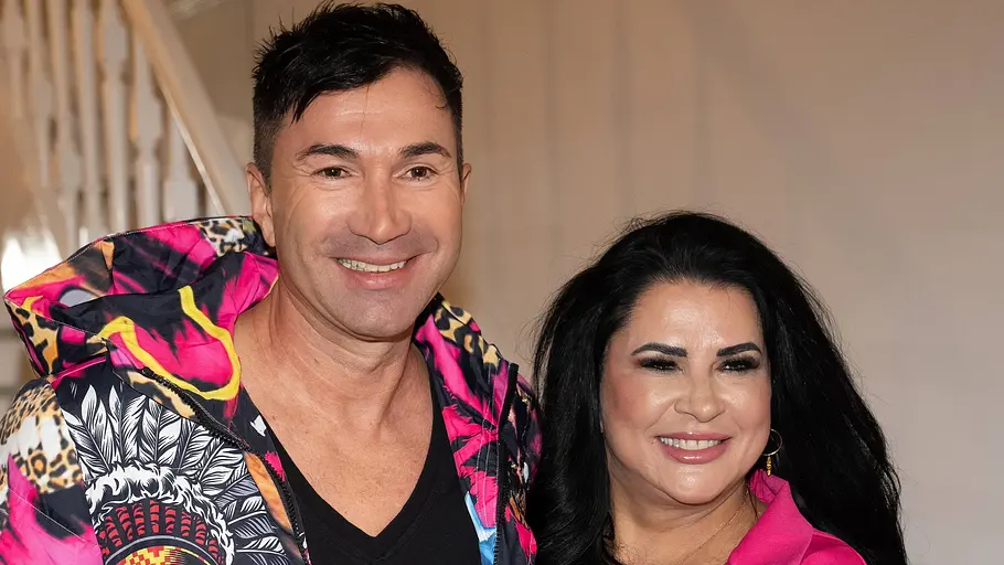 Lucas Cordalis und Iris Katzenberger: Ein Mann in bunt gemusterter Jacke und eine Frau in pinkem Blazer stehen lächelnd nebeneinander vor einer Treppe.