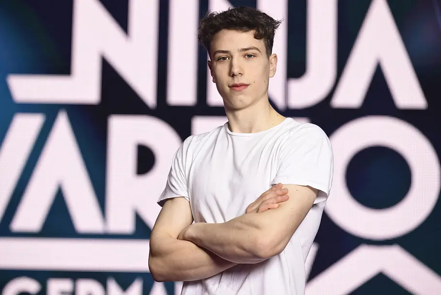 Lukas Homann ist Teilnehmer bei Ninja Warrior Germany 2026 Lukas Homann steht im weißen T-Shirt mit verschränkten Armen vor einem großen „Ninja Warrior“-Schriftzug.