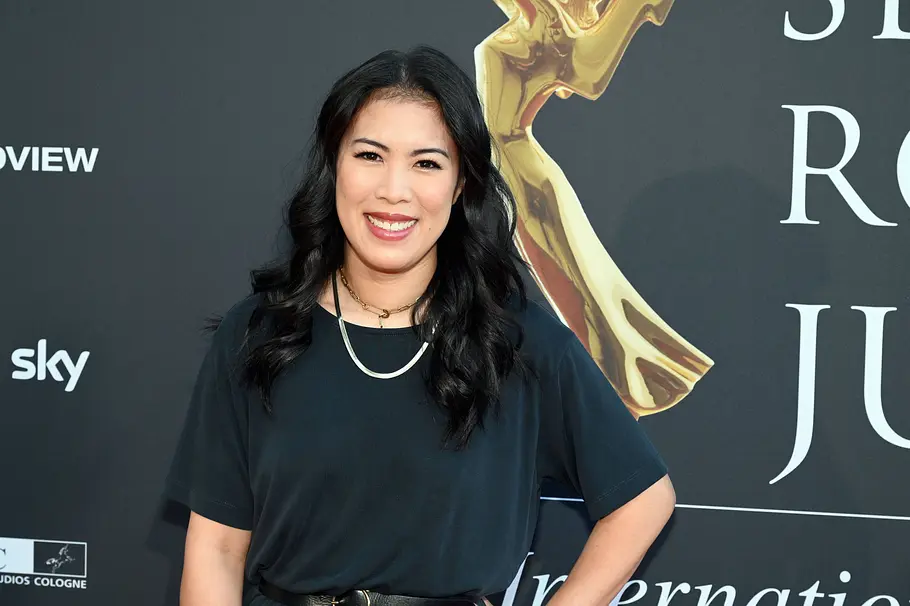 Mai Thi Nguyen-Kim Mai Thi Nguyen-Kim lächelt in schwarzem Oberteil und mit Halsketten vor einer dunklen Wand mit Schriftzügen und goldener Figur.