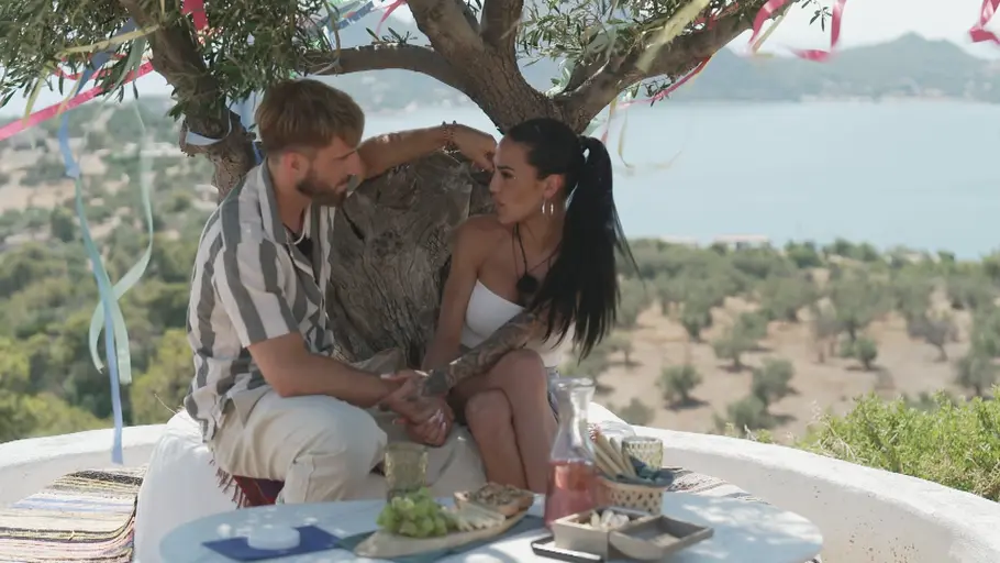 Make Love Fake Love Ein Mann und eine Frau sitzen unter einem mit bunten Bändern geschmückten Baum auf einer Terrasse mit Blick auf Meer und Landschaft, halten sich an den Händen und unterhalten sich; vor ihnen steht ein kleiner Tisch mit Snacks und Getränken.