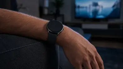 Ein entspannter Arm hängt über der Sofalehne, am Handgelenk eine runde Smartwatch. Im Hintergrund läuft ein Fernseher. - Foto: TV Movie / KI / ChatGPT Ein entspannter Arm hängt über der Sofalehne, am Handgelenk eine runde Smartwatch. Im Hintergrund läuft ein Fernseher. - Foto: TV Movie / KI / ChatGPT