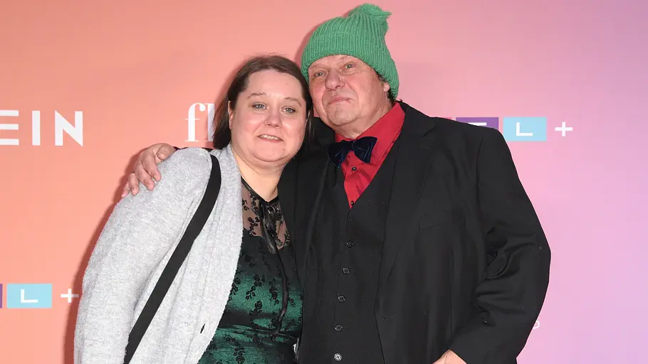 Manni und Jana Ludolf Manni und Jana Ludolf posieren Arm in Arm vor einer rosa Logowand. Er trägt eine grüne Mütze, sie ein grünes Spitzenkleid.