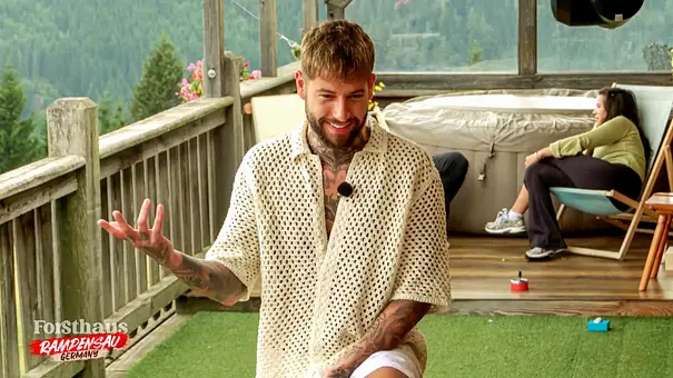 „Reality-TV-Teilnehmer Marc-Robin steht auf der Terrasse des ‚Forsthaus Rampensau‘, gestikuliert mit der Hand und lächelt, während andere Kandidatinnen im Hintergrund sitzen.“ - Foto: Joyn „Reality-TV-Teilnehmer Marc-Robin steht auf der Terrasse des ‚Forsthaus Rampensau‘, gestikuliert mit der Hand und lächelt, während andere Kandidatinnen im Hintergrund sitzen.“ - Foto: Joyn