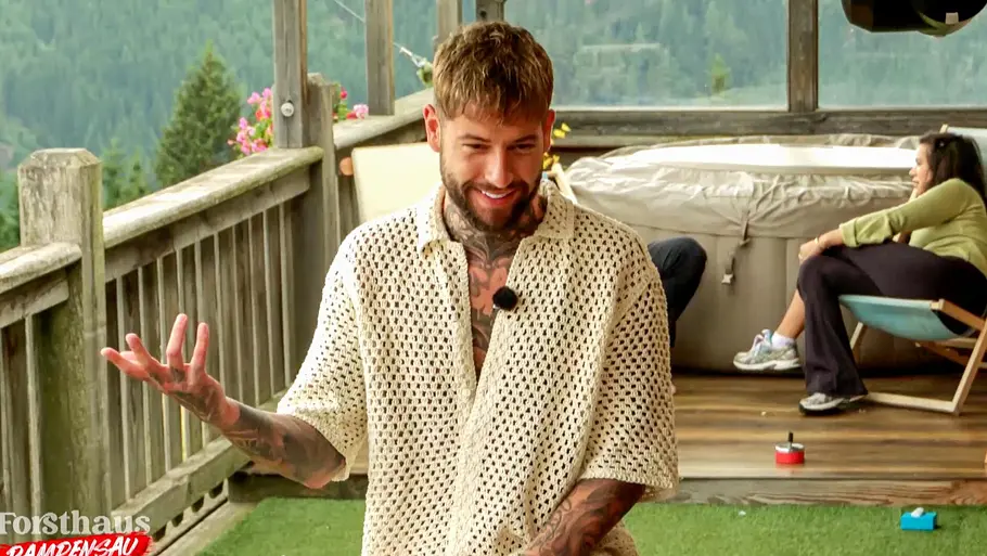 Marc Robin „Reality-TV-Teilnehmer Marc-Robin steht auf der Terrasse des ‚Forsthaus Rampensau‘, gestikuliert mit der Hand und lächelt, während andere Kandidatinnen im Hintergrund sitzen.“