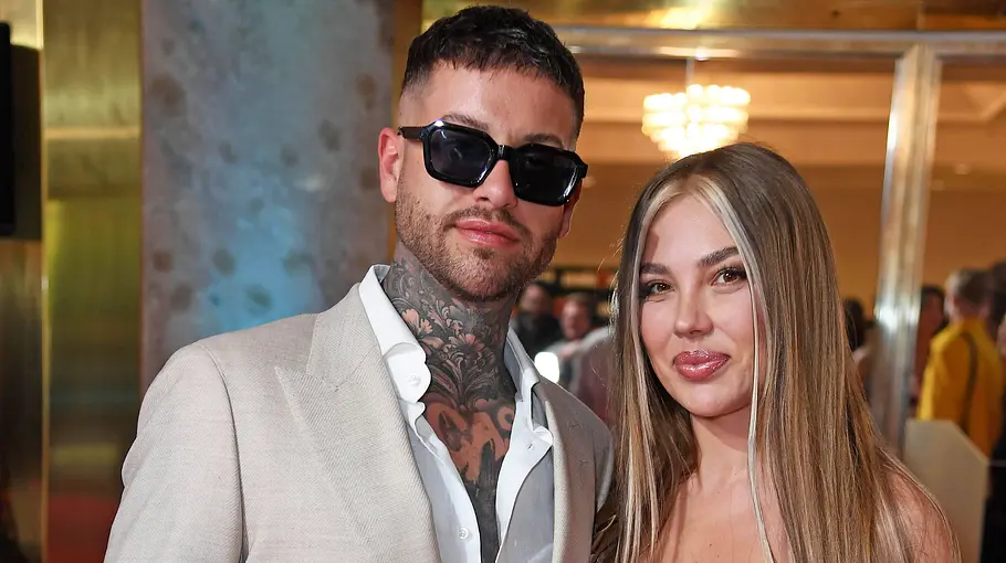 Ina und Marc-Robin posieren auf einem roten Teppich. Er trägt einen hellen Anzug und eine Sonnenbrille. Beide schauen in die Kamera.