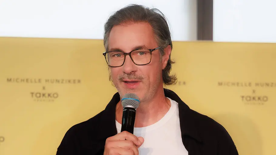 Marco Schreyl trägt Brille, schwarze Jacke und weißes Shirt. Er hält ein Mikrofon und Karten vor einer gelben Werbewand.