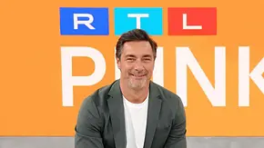 Marco Schreyl sitzt lächelnd in dunklem Sakko und weißem T-Shirt vor einer orangefarbenen Wand mit RTL-Logo. - Foto: RTL / Stefan Gregorowius Marco Schreyl sitzt lächelnd in dunklem Sakko und weißem T-Shirt vor einer orangefarbenen Wand mit RTL-Logo. - Foto: RTL / Stefan Gregorowius