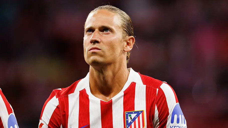 Ein Fußballspieler von Atlético Madrid im rot-weiß gestreiften Trikot blickt vor dem Anpfiff mit ernstem Gesichtsausdruck nach oben. Im Hintergrund ist das Stadion nur unscharf zu erkennen.
