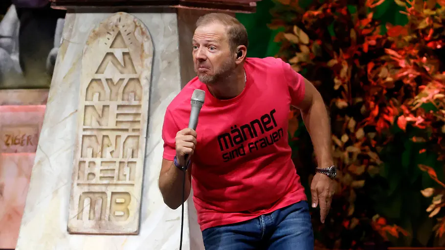 Ein Mann steht auf einer Bühne und hält ein Mikrofon in der Hand. Er trägt ein pinkes T-Shirt mit der Aufschrift „männer sind frauen“ und eine Jeans. In leicht gebückter Haltung schaut er mit ausdrucksstarkem Gesichtsausdruck ins Publikum. Im Hintergrund sind eine marmorne Säule mit eingravierter Schrift sowie dekorative Pflanzen und buntes Bühnenlicht zu sehen.