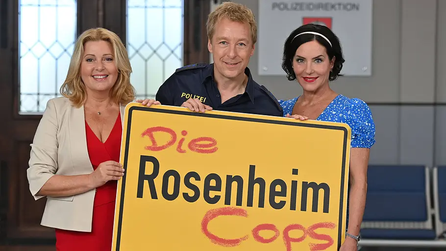 Marisa Burger Karin Thaler, Max Mueller und Marisa Burger halten ein großes Ortseingangsschild mit der Aufschrift „Die Rosenheim Cops vor sich.