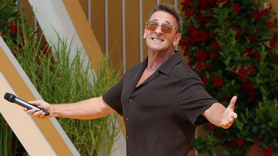 Mark Keller ZDF Weihnachtsshow Mark Keller lächelt mit ausgebreiteten Armen, Sonnenbrille und Mikrofon vor grünen Pflanzen und roten Blüten.