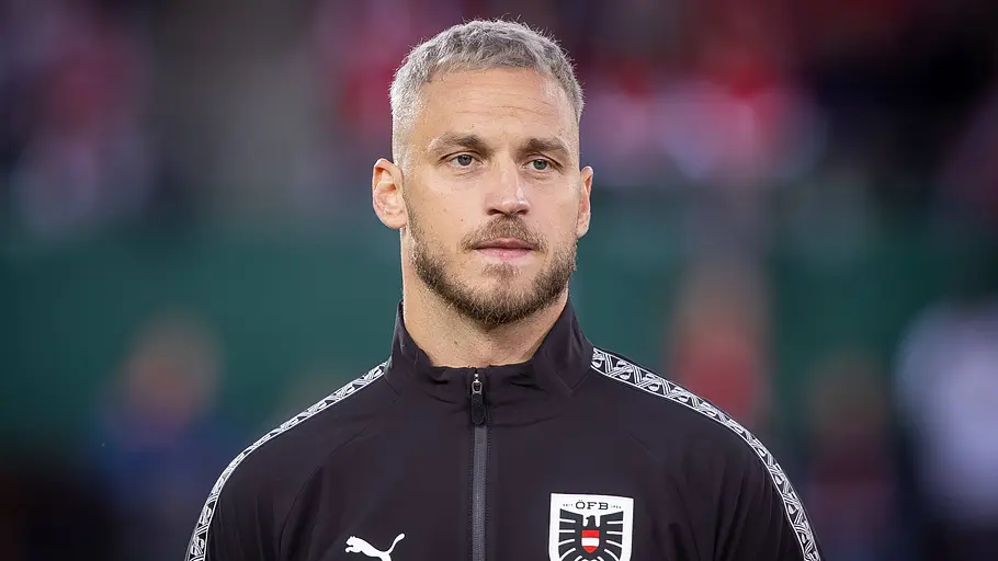 Fußballspieler der österreichischen Nationalmannschaft in schwarzer Trainingsjacke mit ÖFB-Wappen blickt vor einem Länderspiel konzentriert nach vorne. Im Hintergrund sind die verschwommenen Ränge des Stadions zu sehen.