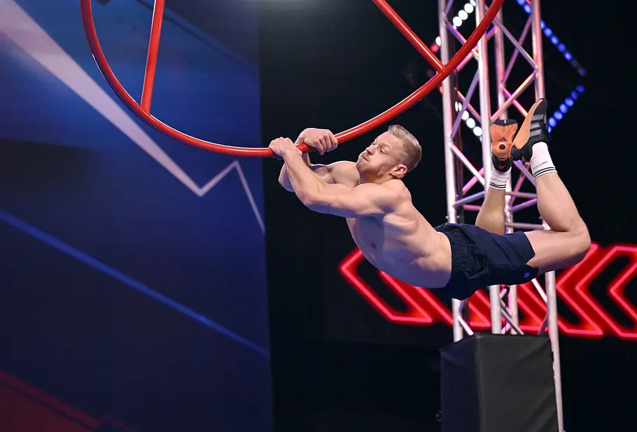 Markus Gebauer ist Teilnehmer bei Ninja Warrior Germany 2026 Markus Gebauer hängt mit freiem Oberkörper und angewinkelten Beinen an einem großen roten Ring im beleuchteten Parcours.