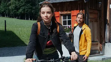 Mia hält im Vordergrund einen Fahrradlenker. Hinter ihr steht Markus in gelber Jacke vor einem Holzhaus mit roten Fensterläden. - Foto: ZDF/Sabine Finger Fotografie Mia hält im Vordergrund einen Fahrradlenker. Hinter ihr steht Markus in gelber Jacke vor einem Holzhaus mit roten Fensterläden. - Foto: ZDF/Sabine Finger Fotografie