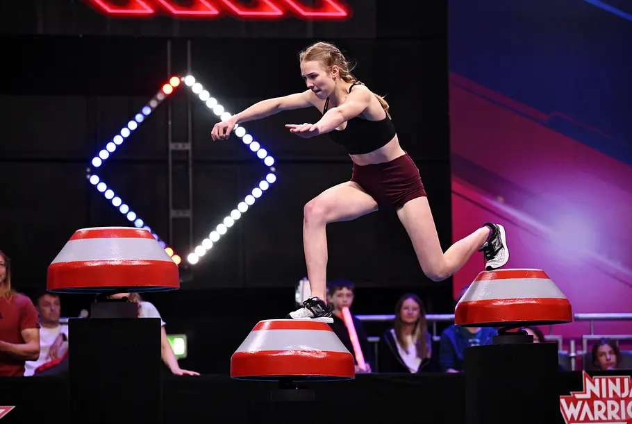 Marlene Kern ist Teilnehmerin bei Ninja Warrior Germany 2026 Marlene Kern springt mit ausgestreckten Armen zwischen rot-weißen Podesten. Sie trägt ein schwarzes Top und bordeauxrote Shorts.