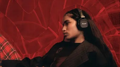 Eine Person mit langen dunklen Haaren trägt schwarze Marshall On-Ear-Kopfhörer und lehnt entspannt vor einem roten, abstrakten Hintergrund. - Foto: Amazon/PR